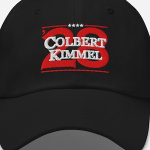 May include: Black baseball cap with the embroidered text "COLBERT KIMMEL 28" in red and white. The cap has a curved brim and a six-panel design. The text is centered on the front of the cap.