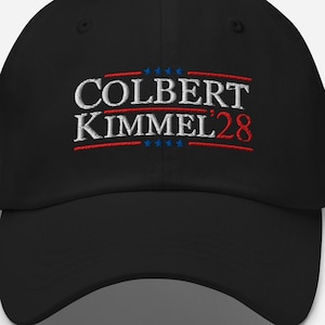 May include: Black baseball cap with the embroidered text "COLBERT KIMMEL 28" in white, red, and blue. The text is arranged in a horizontal format across the front of the cap.