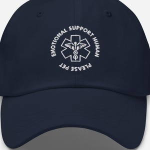 Può includere: Cappellino da baseball blu navy con visiera curva. Il cappellino presenta testo bianco e un simbolo medico che recita "EMOTIONAL SUPPORT HUMAN PLEASE PET". Il cappello è uno stile classico e regolabile.