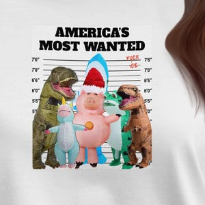 May include: White t-shirt featuring a graphic of cartoon characters in a lineup with the text "AMERICA'S MOST WANTED." The characters include a dinosaur, a pig wearing a Santa hat, and other toy figures.