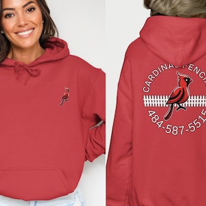 May include: Red hoodie with a small cardinal logo on the front and a larger circular design on the back. The back design features a red cardinal, a white picket fence, and the words "CARDINAL FENCING" and a phone number.