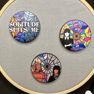 Puede incluir: Tres pines redondos con diseños coloridos. El primer pin tiene un diseño de telaraña azul, rosa y amarillo con el texto "SOLITUDE SUITS ME". El segundo pin tiene un diseño de telaraña colorido con una silueta negra de una persona con un vestido. El tercer pin tiene un diseño de telaraña colorido con una silueta negra de dos personas.
