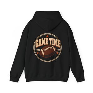 May include: A black hooded sweatshirt featuring a circular graphic on the back. The design includes the text "GAME TIME" in a bold, light-colored font, a brown football illustration, and small orange stars arranged within a circular border.
