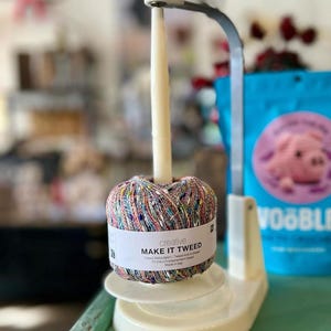 May include: A cream-colored yarn holder with a multi-colored ball of yarn labeled "MAKE IT TWEED." The yarn is held on a vertical spindle. A blue bag with a pig graphic is in the background.