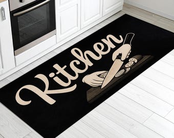 May include: Black rectangular kitchen rug with the word "Kitchen" in a cursive font. The rug features a graphic of a hand holding a knife, slicing vegetables on a cutting board. The rug is placed in front of a white kitchen cabinet.