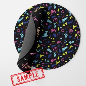 May include: A round black mouse pad with a colorful geometric pattern. The pattern includes pink, blue, yellow, and green lines and shapes on a black background. The mouse pad is rolled up and has a "SAMPLE" stamp on it.