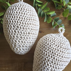 May include: Two cream-colored crocheted egg-shaped ornaments with a loop at the top. The ornaments are on a wooden surface with green leafy branches.