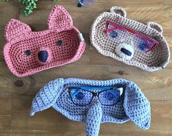 May include: Three crocheted animal-shaped eyeglass holders. A pink fox, a beige bear, and a gray elephant. Each holder has a pair of glasses resting inside.
