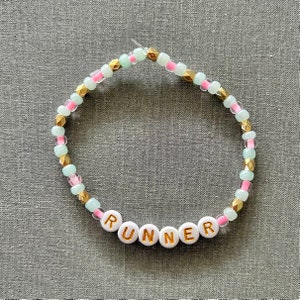May include: A beaded bracelet with alternating light green and pink beads and gold accents. The bracelet spells out the word "RUNNER" in white beads with gold lettering.