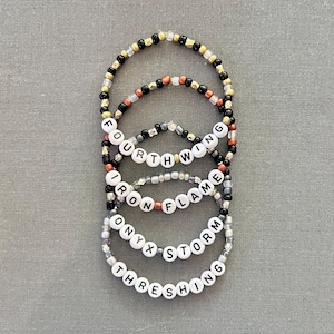 May include: A set of four beaded bracelets featuring white letter beads spelling out FOURTH WING, IRON FLAME, ONYX STORM, and THRESHING. The bracelets are constructed with a mix of black, gold, copper, silver, and clear glass beads in various sizes.