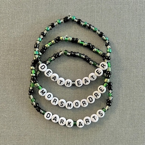 May include: Four layered beaded bracelets with green and black beads. The bracelets spell out "Dark Arts", "Morsmordre", "Death Eater", and "Always".