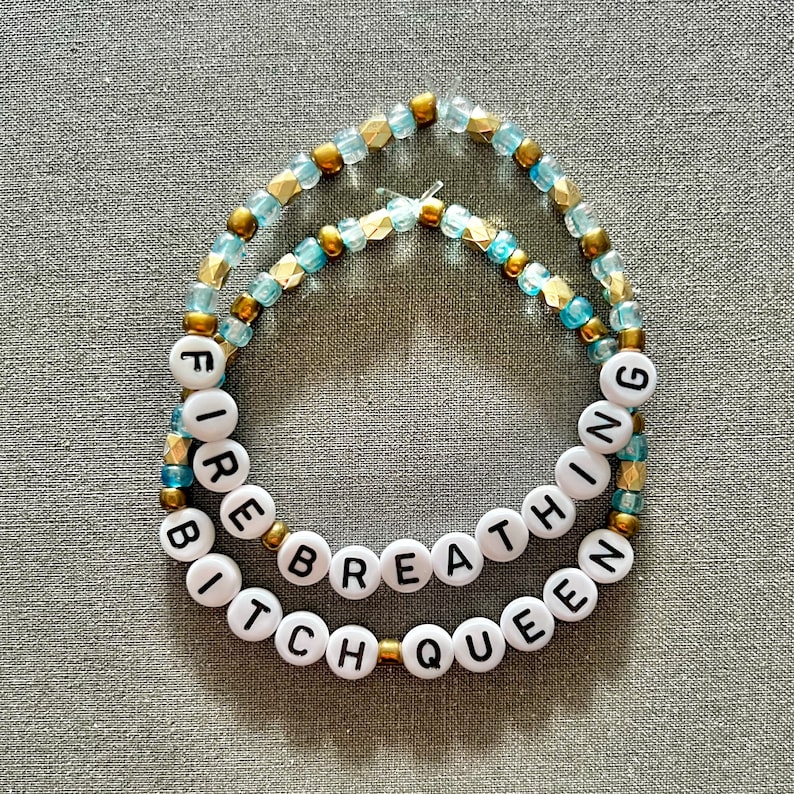May include: Two beaded bracelets with the words "Fire Breathing Bitch Queen" spelled out in black letters on white beads. The beads are strung on gold and blue thread.