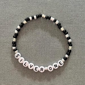 May include: A black and silver beaded bracelet with the word "SILVER ONE" spelled out in white beads.