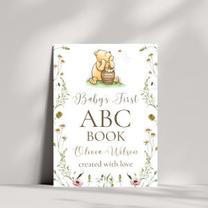 K&ouml;nnte beinhalten: Eine wei&szlig;e Karte mit einer Winnie-the-Pooh-Illustration und gr&uuml;nem floralen Rand. Der Text lautet "Baby's First ABC Book Olivia Wilson created with love."