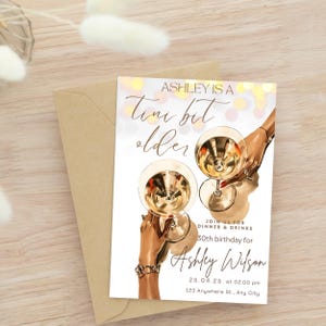 May include: A white invitation card with gold text and a graphic of two hands holding champagne flutes. The text reads "Ashley is a tiny bit older" and includes details for a 30th birthday party.