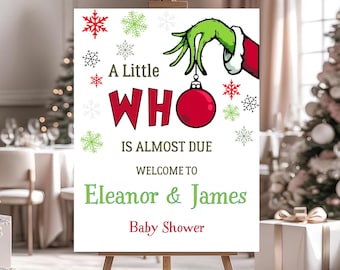 May include: A white sign on a wooden easel with the text "A Little WHO is Almost Due" in red and green. The sign is decorated with snowflakes and a Grinch-themed hand holding a red ornament. The sign welcomes guests to Eleanor & James' baby shower.