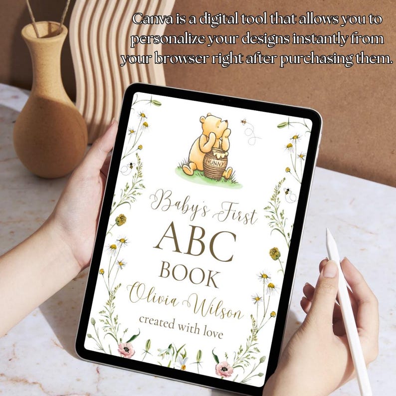 K&ouml;nnte beinhalten: Ein digitales Tablet zeigt ein ABC-Buch-Design f&uuml;r Babys mit Winnie Puuh. Das Design enth&auml;lt den Text "Baby's First ABC Book" und "Olivia Wilson created with love". Das Bild ist von Blumen- und Bienenillustrationen umgeben.