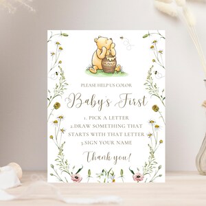 K&ouml;nnte beinhalten: Rechteckiges Schild mit einer Illustration von Winnie Puuh, einem Honigtopf und einem floralen Rand. Auf dem Schild steht "Baby's First" und es enth&auml;lt Anweisungen zum Ausw&auml;hlen eines Buchstabens, Zeichnen und Unterschreiben des Namens. Das Design umfasst Bienen und Blumen.