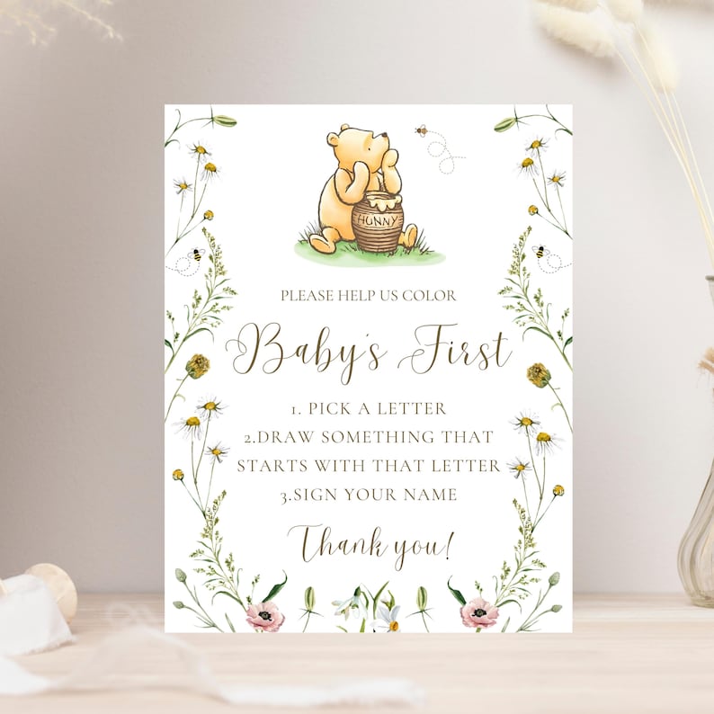 K&ouml;nnte beinhalten: Rechteckiges Schild mit einer Illustration von Winnie Puuh, einem Honigtopf und einem floralen Rand. Auf dem Schild steht "Baby's First" und es enth&auml;lt Anweisungen zum Ausw&auml;hlen eines Buchstabens, Zeichnen und Unterschreiben des Namens. Das Design umfasst Bienen und Blumen.