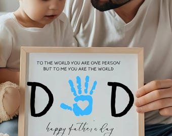 May include: A white framed sign with the text "To the world you are one person but to me you are the world" and a blue handprint with a heart in the center. The sign also says "DD happy father's day 2025".
