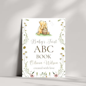 K&ouml;nnte beinhalten: Rechteckiges Schild mit einer Aquarellillustration von Winnie Puuh und einem Honigtopf. Das Schild tr&auml;gt die Aufschrift "Baby's First ABC Book" mit dem Namen "Olivia Wilson" und "created with love", umgeben von floralen Elementen.