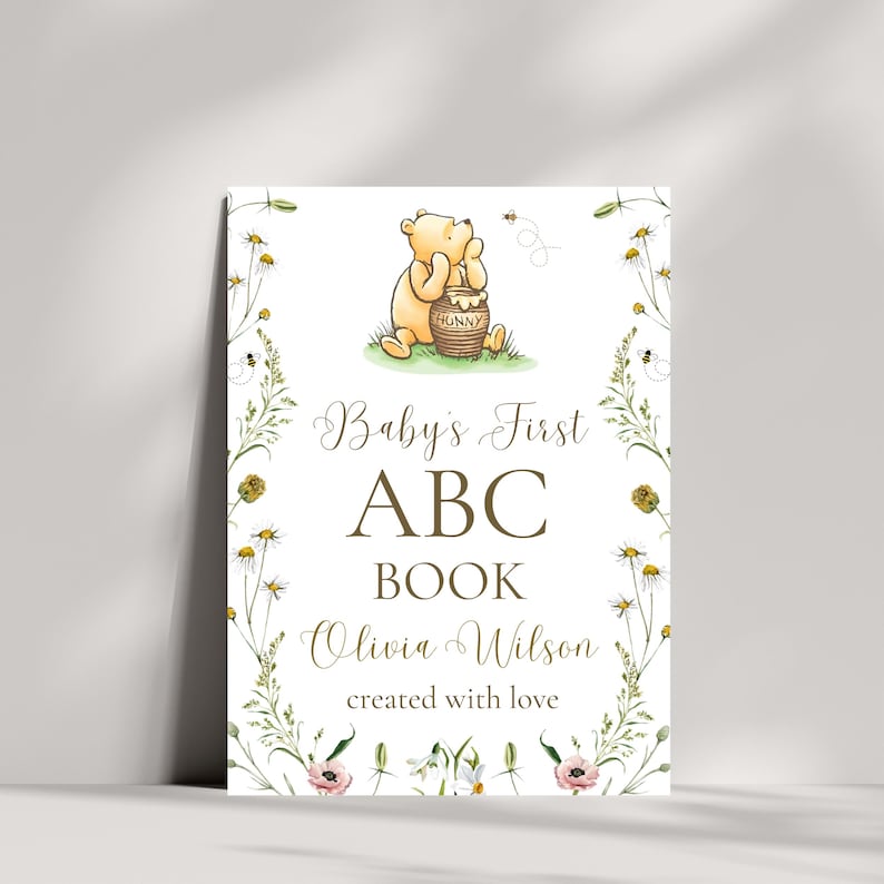 K&ouml;nnte beinhalten: Rechteckiges Schild mit einer Aquarellillustration von Winnie Puuh und einem Honigtopf. Das Schild tr&auml;gt die Aufschrift "Baby's First ABC Book" mit dem Namen "Olivia Wilson" und "created with love", umgeben von floralen Elementen.