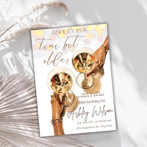 May include: A white invitation card with a gold and pink background. The text reads "Ashley is a Tini bit older" and includes details for a 30th birthday party. The card features a hand holding a martini glass.