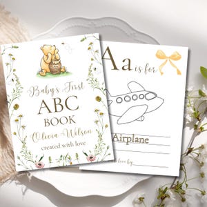 K&ouml;nnte beinhalten: Zweiseitiges Baby-ABC-Buch. Das Cover zeigt Winnie Puuh mit einem Honigtopf, den Text "Baby's First ABC Book" und den Namen "Olivia Wilson". Die zweite Seite hat den Buchstaben "A" und eine Strichzeichnung eines Flugzeugs.