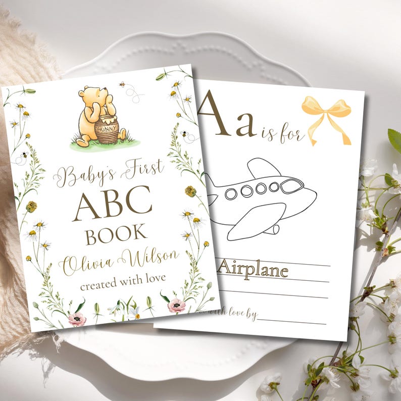 K&ouml;nnte beinhalten: Zweiseitiges Baby-ABC-Buch. Das Cover zeigt Winnie Puuh mit einem Honigtopf, den Text "Baby's First ABC Book" und den Namen "Olivia Wilson". Die zweite Seite hat den Buchstaben "A" und eine Strichzeichnung eines Flugzeugs.