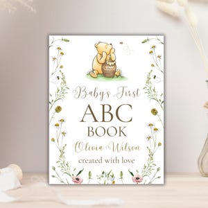 K&ouml;nnte beinhalten: Ein wei&szlig;er Hintergrund mit einer Illustration von Winnie Puuh, der einen Honigtopf h&auml;lt. Der Text lautet "Baby's First ABC Book Olivia Wilson created with love".