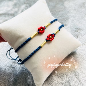 Op de afbeelding: Twee handgemaakte blauwe macramé armbanden met rode bloemaccenten en goudkleurige banden. De armbanden worden gepresenteerd op een wit kussen, waarbij de ingewikkelde knopen en levendige kleuren worden getoond. Een uniek accessoire.
