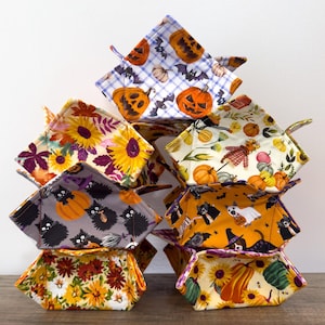 May include: A stack of fabric bowl cozies with various Halloween and fall-themed patterns. Designs include pumpkins, bats, sunflowers, scarecrows, and black cats. The cozies are square with a folded design and are stacked on top of each other.