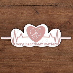 Puede incluir: Una pegatina con un corazón rosa y una silueta de bebé, sobre un fondo blanco. El diseño incluye una línea de latido cardíaco y el texto "Every heartbeat matters". La pegatina está sobre una superficie de madera.