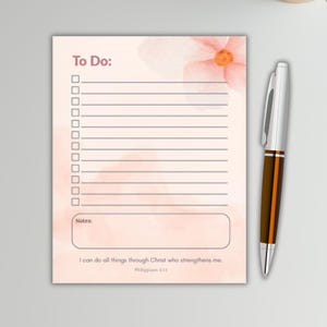 Op de afbeelding: Een notitieblok met een bloemenontwerp en de woorden "To Do:" bovenaan. Het notitieblok heeft lijnen om op te schrijven en een sectie voor notities. Een pen met een zilveren en bruine schacht ligt naast het notitieblok. De tekst "I can do all things through Christ who strengthens me. Philippians 4:13" staat onderaan.