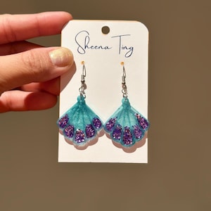 Op de afbeelding: Een paar turquoise en paarse glitter schelpoorbellen. De oorbellen zitten op een wit label met de handgeschreven tekst "Sheena Ting". De oorbellen hebben zilverkleurige haken.