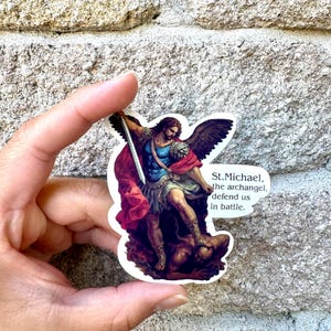 May include: A die-cut sticker featuring a depiction of Saint Michael the Archangel with dark wings, wearing blue and red armour, holding a sword, and standing over a figure. The text reads: St. Michael, the archangel, defend us in battle.