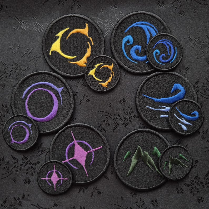 May include: A collection of embroidered patches in various designs. Each patch is circular with a black background and features colorful abstract symbols in yellow, blue, purple, green, and pink. The patches vary in size, with some larger and some smaller.