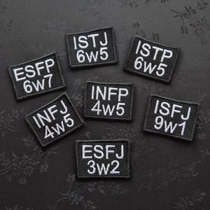 Può includere: Una collezione di sette toppe nere ricamate con testo bianco. Ogni toppa mostra un'abbreviazione del tipo di personalità di quattro lettere e una combinazione di numeri. Le toppe sono disposte su una superficie scura e strutturata.