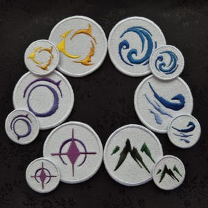 May include: A collection of embroidered patches with various elemental symbols. The patches are circular, with white fabric and detailed designs in yellow, blue, purple, and green. The designs include stylized waves, mountains, and abstract shapes.