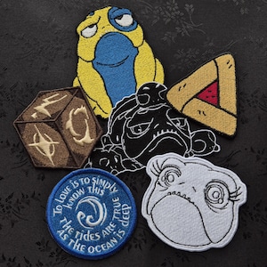 May include: A selection of embroidered patches with diverse designs. These include a yellow and blue frog, a black and white frog, a brown die, a blue patch with text, and a triangle with a red centre. The text on the blue patch reads "To love is to simply know this, as the tides are true, the ocean is deep."