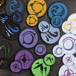 May include: A collection of embroidered patches in various sizes and colors. Designs include abstract shapes, swirls, and geometric patterns in shades of blue, yellow, purple, green, and white. The patches are circular and have a stitched border.