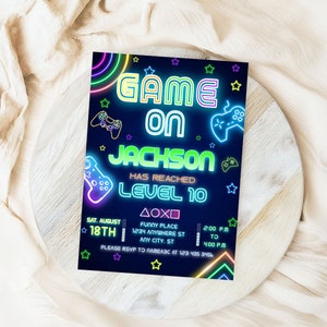 以下が含まれることがあります： 暗い青色の背景にネオン風の招待状。「GAME ON JACKSON HAS REACHED LEVEL 10」のテキストは、ネオンの緑と青のアクセントで表示されます。コントローラーのグラフィックとイベントの詳細が含まれています。