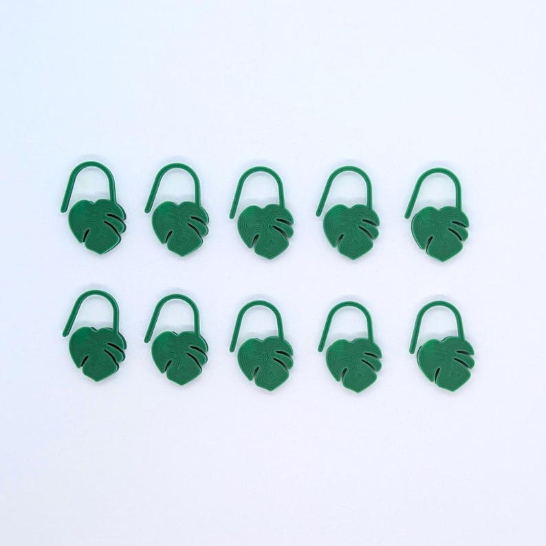 Puede incluir: Diez clips de papel verdes con forma de hoja sobre un fondo blanco. Cada clip presenta un dise&ntilde;o de hoja de monstera con una parte superior curvada. Los clips est&aacute;n espaciados uniformemente.