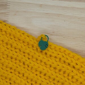 Puede incluir: Primer plano de una tela de ganchillo amarilla brillante con un marcador de puntada verde en forma de hoja adjunto. La tela tiene un aspecto texturizado. El marcador de puntada es de color verde vibrante.