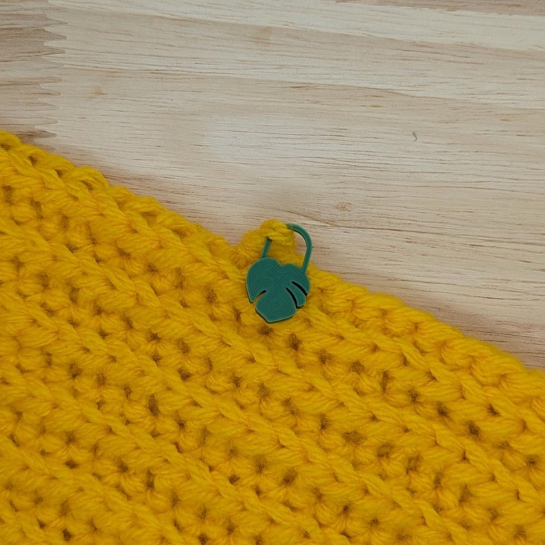 Puede incluir: Primer plano de una tela de ganchillo amarilla brillante con un marcador de puntada verde en forma de hoja adjunto. La tela tiene un aspecto texturizado. El marcador de puntada es de color verde vibrante.