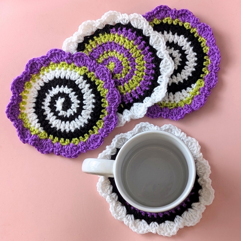May include: A collection of handmade crochet coasters in a variety of colors. The coasters feature a spiral design with black, white, purple, and green accents. One coaster holds a white mug. The coasters have a scalloped edge.