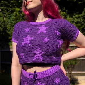 May include: A two-piece crochet set in shades of purple. The top is a short-sleeved crop top with a crew neck, and the bottoms are high-waisted shorts. Both pieces feature a repeating star pattern in a lighter shade of purple.