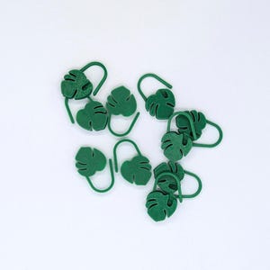 Puede incluir: Un conjunto de marcadores de plantas verdes con forma de hoja. Cada marcador presenta un dise&ntilde;o de hoja de monstera con un gancho curvado para facilitar su colocaci&oacute;n en el suelo. Los marcadores son de material verde liso sobre fondo blanco.