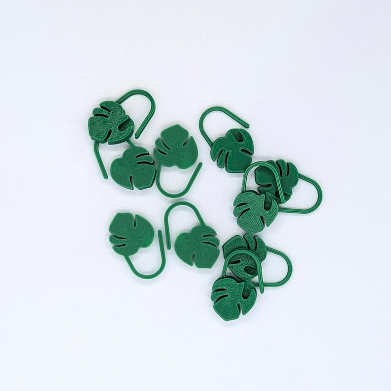 Puede incluir: Un conjunto de marcadores de plantas verdes con forma de hoja. Cada marcador presenta un dise&ntilde;o de hoja de monstera con un gancho curvado para facilitar su colocaci&oacute;n en el suelo. Los marcadores son de material verde liso sobre fondo blanco.