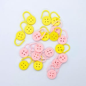 May include: A collection of yellow and pink stitch markers, each with a button-like design and a curved hook. The markers are scattered against a white background, ideal for knitting and crochet projects.